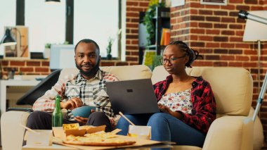 Young people in relationship watching tv show at home, eating snacks and using laptop to browse online app. Man and woman having fun with movie or film, relaxing at television. Tripod shot.