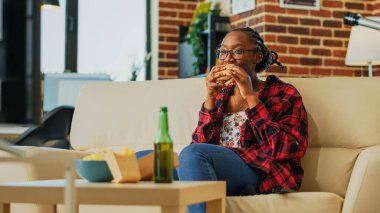 Young woman eating cheeseburger with fries and beer, binge watching favorite tv show by herself at home. Modern girl feeling happy with fast food takeaway, watch film on television. Handheld shot.