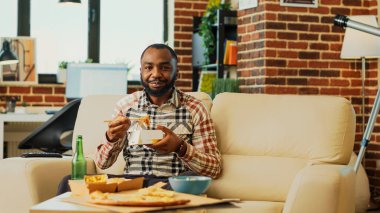 Relaxed young man using chopsticks to eat noodles, having fun watching movie on television. Cheerful person enjoying asian fast food delivery at home, looking at tv show. Tripod shot.