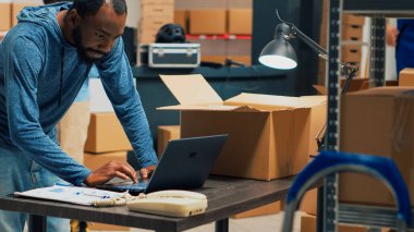 African american man putting products in cardboard boxes, working on distribution and order shipping. Young person checking merchandise and sending supplies, business development.
