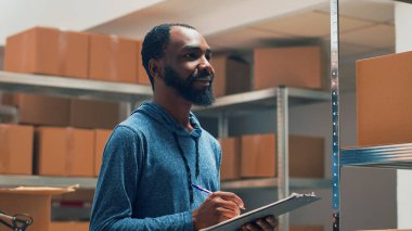 African american owner working on stock inventory, taking notes on clipboard files. Young male employee checking supplies in cardboard boxes, looking at goods on storehouse shelves. Handheld shot.