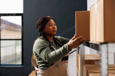 Depo müdürü karton kutularla çalışıyor, depodaki müşteriler için paketler hazırlıyor. Endüstriyel giyinen Afro-Amerikan çalışanlar depoda nakliye ürünlerinde çalışıyorlar.