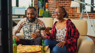 Boyfriend and girlfriend enjoying pizza slices and binge watching show in living room, eating fast food meal. Young couple receiving takeout food order and sitting on couch to have dinner.