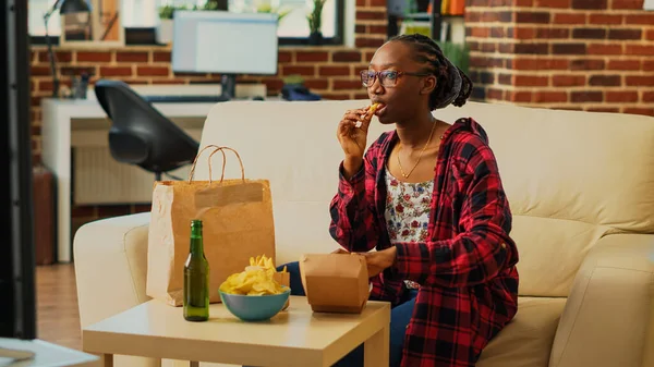 Happy woman eating hamburgers with fries and beer, having fun with action film on television. Smiling person enjoying burger and snacks in bowl, binge watching tv show in living room.