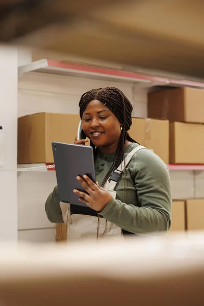 African american employee discussing products inventory with remote manager using landline phone, checking customer online order on tablet computer. Supervisor working in storehouse