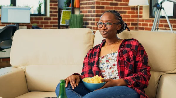 Happy woman laughing at comedy show on television, eating chips and snacks from bowl in living room. Smiling modern girl serving multiple fast food meals from takeout, drinking beer.