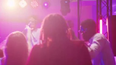 Erkek DJ kulüpte insanlarla parti yapıyor, diskotekte eğlenceli parti atmosferi yaratmak için ses istasyonunda müzik karıştırıyor. Genç adam dans ediyor ve arkadaşlarıyla zıplıyor, dans pistinde eğleniyor..