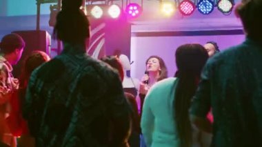 Partide karaoke yapan mutlu bir kadın dans pistinde arkadaşlarının önünde DJ 'le şarkı söylüyor. Kendine güvenen kız, gece kulübündeki elektronik karıştırma istasyonunun yanında canlı şov perfromantizmiyle eğleniyor..