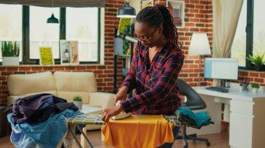 African american housewife using iron on ironing board to do housekeeping work, casual adult ironing clothes in living room. Young woman smoothing out garment and clothing, chores.