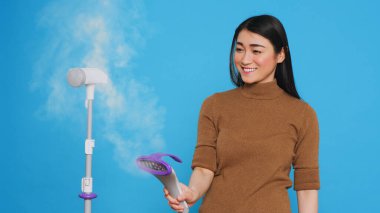 Smiling cleaning lady using steamer to ironing clothes in studio over blue background. Woman used the best cleaning products and equipment to ensure a sparkling, fresh-smelling home for her clients.