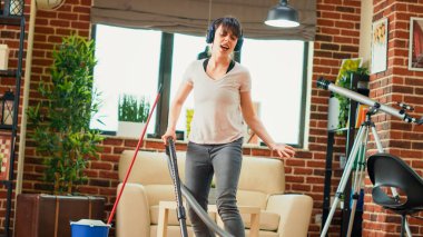 Mutlu ev hanımı kulaklıkla müzik dinliyor, yerleri süpürüyor, elektrik süpürgesiyle eğleniyor. Evde bahar temizliği yaparken gülümseyen genç insan pozitif hissediyor. El kamerasıyla..