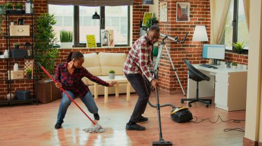 African american couple dancing and cleaning apartment rooms, using mop to wash dirt and vacuum to clean floors. Young life partners enjoying spring cleaning with washing solution.