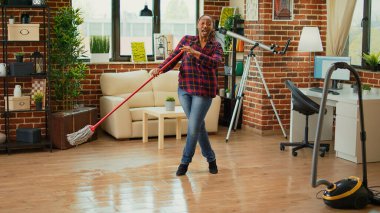 Modern housewife doing dance moves in living room and mopping wooden floors, listening to music and enjoying spring cleaning. Happy person singing and using mop to clean at home.