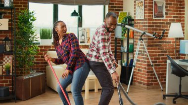 Cheerful people showing dance moves and doing spring cleaning, having fun with music while they clean wooden floors with mop and vacuum cleaner. Young couple smiling and doing chores.