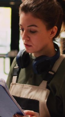Dikey video: Envanter listesi üzerinde çalışan genç yetişkin, depolama odasındaki ürünleri kontrol ediyor. Kargo ve malları inceleyen kadın çalışanlar, lojistik belgeleriyle teftiş yapıyorlar..