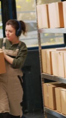 Dikey video: Denetmen raflardaki kutuları düzenliyor, perakende sevkiyatına hazırlanmadan önce paketleri taşıyor. Depoda müzik dinleyen genç bir yetişkin var. El bilgisayarı