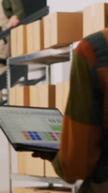 Dikey video: Lojistik teftişi yapmak için dizüstü bilgisayarla çalışan Afrikalı Amerikalı adam, kargo stoğunu doğruluyor. Perakende mağaza dağıtımı için malları inceleyen erkek yönetici, bilgisayardaki ürünleri gözden geçiriyor.