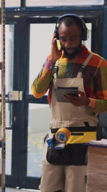 Dikey video: Delikanlı depoda müzik dinlerken envanter sayımı yaparken, raflarda ürünleri incelerken eğleniyor. Erkek depo çalışanı dans ediyor ve kargoya bakıyor, üretim stoku.