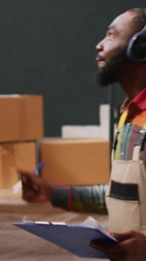Dikey video: Depoda müzik dinleyen işçiler perakende teslimat için raflardaki malları kontrol etmeye çalışıyorlar. Erkek müdür depoda, envanter listesinde dans ederken eğleniyor..