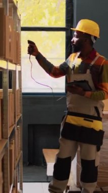 Dikey video: Afrikalı Amerikalı adam depoda barkodları kontrol ediyor, tarayıcı ve tabletle lojistik üzerinde çalışıyor. Raflardaki kutular ile ürün kalite denetimi gözden geçiriliyor, ikmal denetimi yapılıyor.