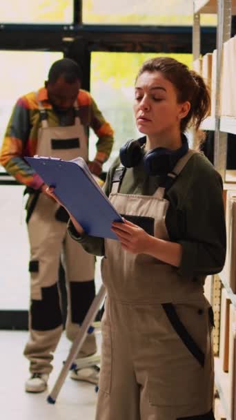 垂直录像 女仓库工人在储藏室检查库存清单 分析装满纸板箱的架子 妇女穿着工作服检查文件上的商品库存 质量控制 — 图库视频影像