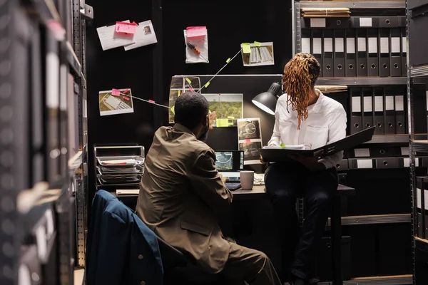 侦探搭档分析监视照片挂在证物板上工作到很晚研究犯罪现场照片和法医报告的非裔美国男女警察 — 图库照片