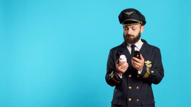 Male aviator looking at bottles of pills in studio, feeling unsure about medicaments and medical treatment. Young adult flying pilot holding jars of medicine and supplements, aviation worker.