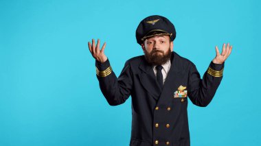 Clueless flying pilot doing i dont know gesture, acting uncertain and doubtful over blue background. Professional aviator feeling unsure and raising shoulders on camera, aircraft captain.