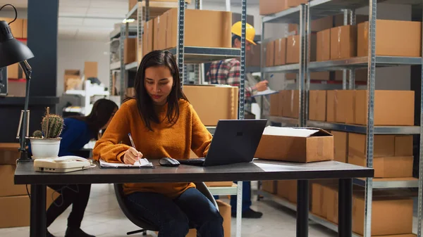 Young person doing stock inventory with laptop, writing logistics information on papers for business development. Female employee planning products distribution in warehouse space.