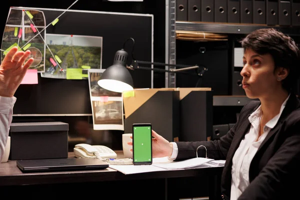 手持绿色显示屏手机的警察模拟展示 与警察讨论犯罪学报告 调查员在黑暗的房间里加班 带有独立模板的电话 — 图库照片