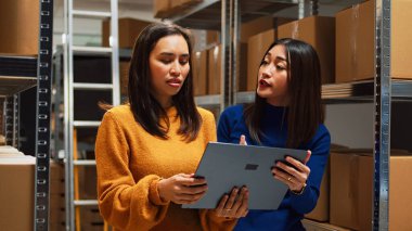 Management team arranging goods on shelves, using laptop to plan stock logistics in warehouse space. Female employees looking at packs of retail products on racks, business plan.