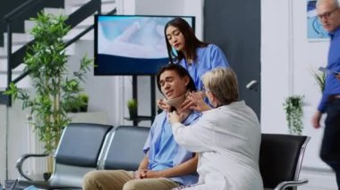 Hastanenin bekleme salonunda hastaya boyun tasması takılmasına yardım eden yaşlı doktor, sağlık tedavisini açıklıyor. Travmatolog, sağlık kontrolünde destek alıyor.