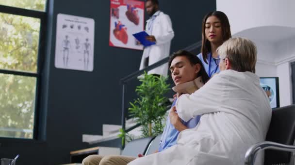 病院の待合室での診察中に負傷者が子宮頸部を取るのを助けるシニア医師 首の損傷後のアジア人男性と医療専門知識を議論する医療スタッフ — ストック動画