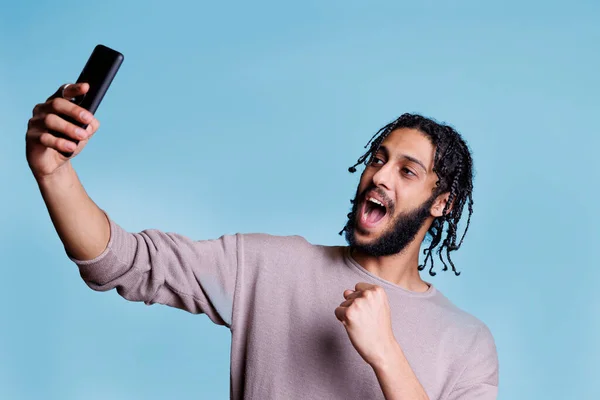 兴奋的Arab男人在智能手机上拿着自己的东西 同时握紧拳头 表现出胜利的姿态 年轻而快乐的人摆出一副快乐的样子 摆出一副手机照片 — 图库照片