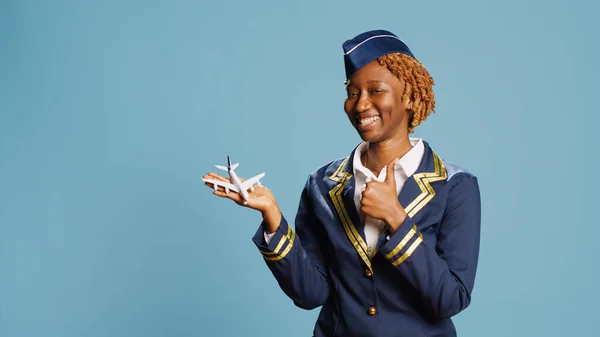 Smiling air hostess holding fake small airplane, playing with artificial miniature plane toy in studio. Cheerful happy airliner having fun with mini imitation aircraft on blue background.