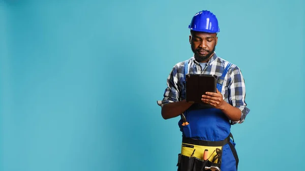 Young handyman using digital tablet on camera, preparing to start constuction contract. Male carpenter in uniform browisng online social media page on modern gadget, building site.