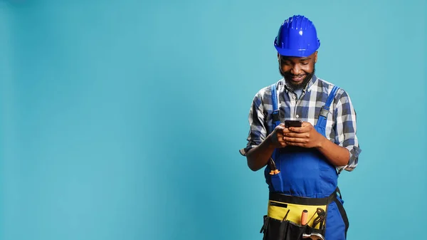 Construction worker texting messages on smartphone app, using social media browser in studio. Young craftsman browsing online website, wearing building overalls and safety helmet.