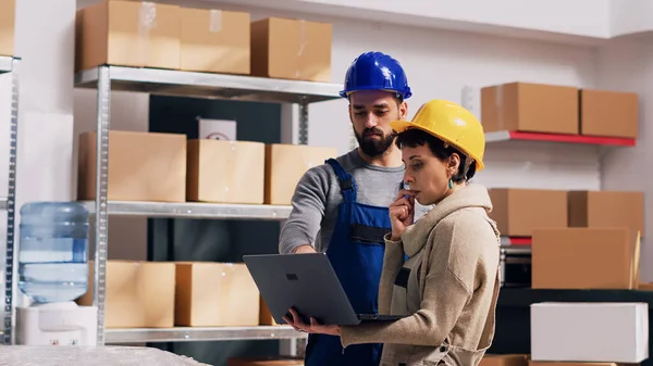Diverse employees talking about stock logistics in depot, merchandise in boxes placed on storage room racks. Man and woman wearing overalls and hardhats using laptop in warehouse space.