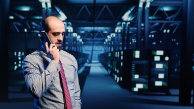 Bilgi güvenliği görevlisi telefonla konuşuyor, büyük veri ve sunucu raflarıyla profesyonel veri merkezinde çalışıyor. Bilgi işlem teknisyeni akıllı telefon kullanıyor, ağ sistemini inceliyor. Üçayak atışı.