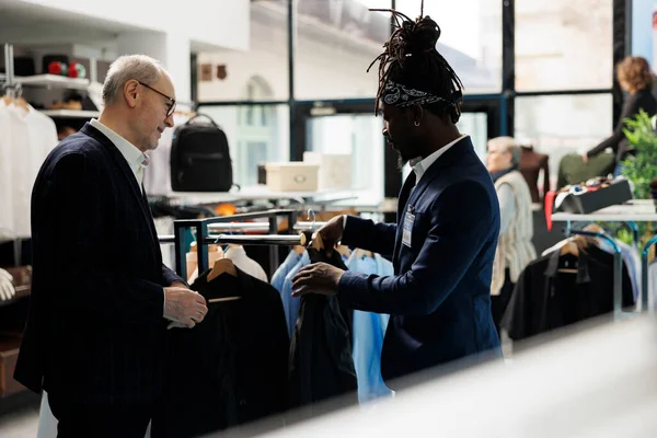 アフリカ系アメリカ人の従業員は ショッピングセンターでスーツで顧客を支援し クライアントは現代的な服を購入しようとしています ショールームでファッショナブルな商品を購入する高齢者 ファッションコンセプト — ストック写真