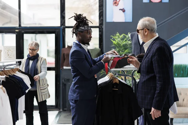 アフリカ系アメリカ人従業員は 現代のブティックで生地を分析し 適切なネクタイで顧客を支援します ショールームでカジュアルウェアやファッショナブルな商品のためのスタイリッシュなスーツショッピングを身に着けている高齢者 — ストック写真