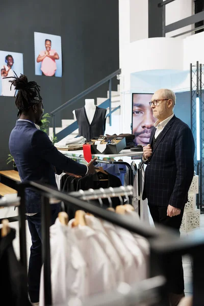 Senior client wanting to buy formal suit in clothing store, asking showroom worker for help to choose the right clothes. Elderly man buying fashionable merchandise and trendy accessories