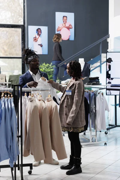 ブティックワーカーは 商業活動中に白いシャツを選ぶことでスタイリッシュな顧客を支援します アフリカ系アメリカ人の顧客は ショッピングセンターでファッショナブルな服やアクセサリーを購入 ファッションコンセプト — ストック写真