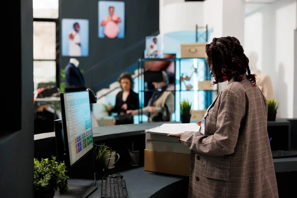 非洲裔美国人雇员站在收银机旁 在购物中心的计算机上检查商品库存 营业员为顾客准备装有时髦服装的套件 — 图库照片