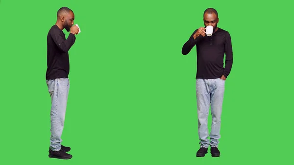 若い人は スタジオでカフェイン飲料のカップを提供し 全身の緑の画面上のコーヒーを飲む ジーンズ姿のカメラでリフレッシュドリンクを楽しむ自信に満ちた男 レジャー活動 — ストック写真