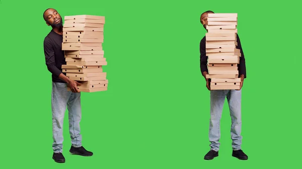 若い人がカメラにピザの大きな山を提供し ファーストフードの注文で多くの段ボール箱を運んでいます 男性大人とともにパッケージ上のフルボディ緑の画面の背景 — ストック写真