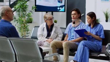 Kıdemli doktor hastane bekleme salonunda hemşireyle tıbbi uzmanlığı tartışıyor ve yaralı hastaya sağlık tedavisini açıklıyor. Muayene sırasında boyunluk takan Asyalı bir adam.