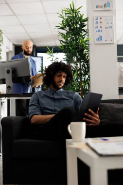 Unicron şirketinin profesyonel elemanı, yeni ofis molasında dijital tablo kullanıyor. Girişimci, modern çalışma ortamında koltukta otururken taşınabilir cihazlarda video izliyor.