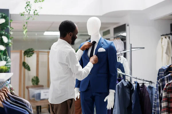 Clothing store stylist creating formal wear outfit on mannequin in retail outlet. African american man dressing dummy model in suit and fixing tie while working in shopping mall