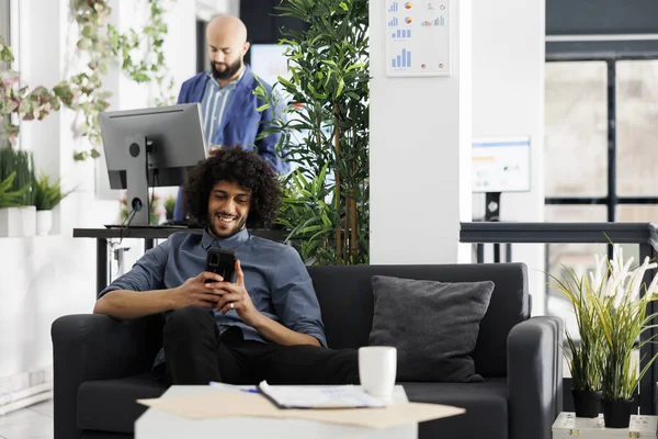 兴高采烈的阿拉伯企业家一边坐在沙发上创业 一边用智能手机在线聊天 在合作空间看手机搞笑视频的商人大笑 — 图库照片